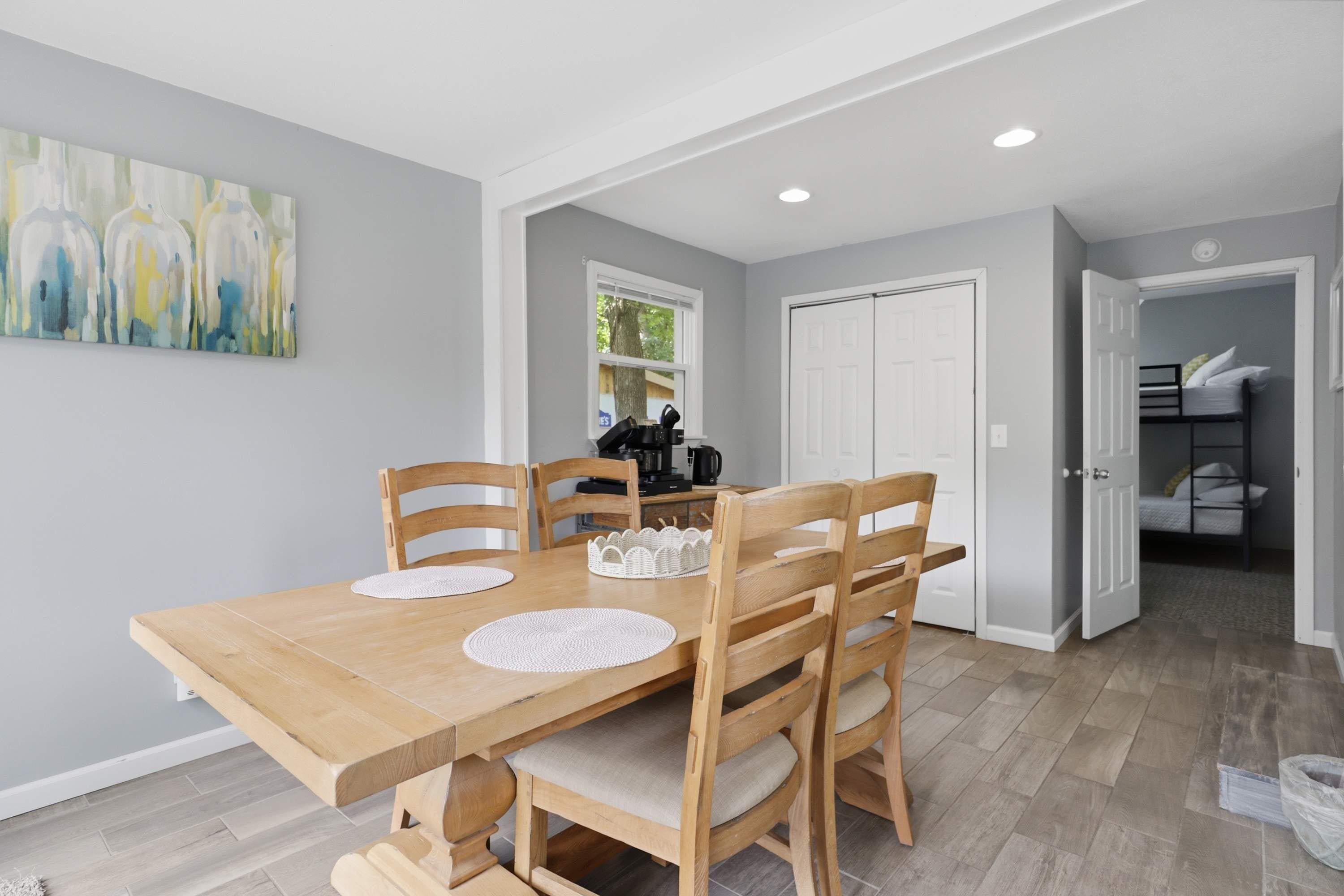 A dining area with a wooden table and chairs, a painting on the wall, and a kitchen in the background.