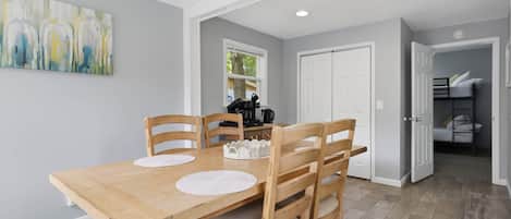 A dining area with a wooden table and chairs, a painting on the wall, and a kitchen in the background.
