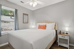 Un dormitorio con una cama, una mesita de noche, un ventilador de techo, una ventana con cortinas y un espejo en la pared.