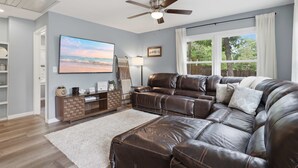 A modern living room with a sectional sofa, a flat-screen TV, a ceiling fan, and a large window with curtains. - Lakefront Home 3 Bedroom 2 Bath With Private Dock (Gravois Mills)