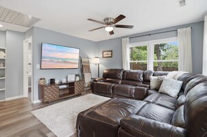 A modern living room with a sectional sofa, a flat-screen TV, a ceiling fan, and a large window with curtains.