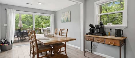 A dining area with a wooden table and chairs, a coffee station, and a view of greenery through large windows.