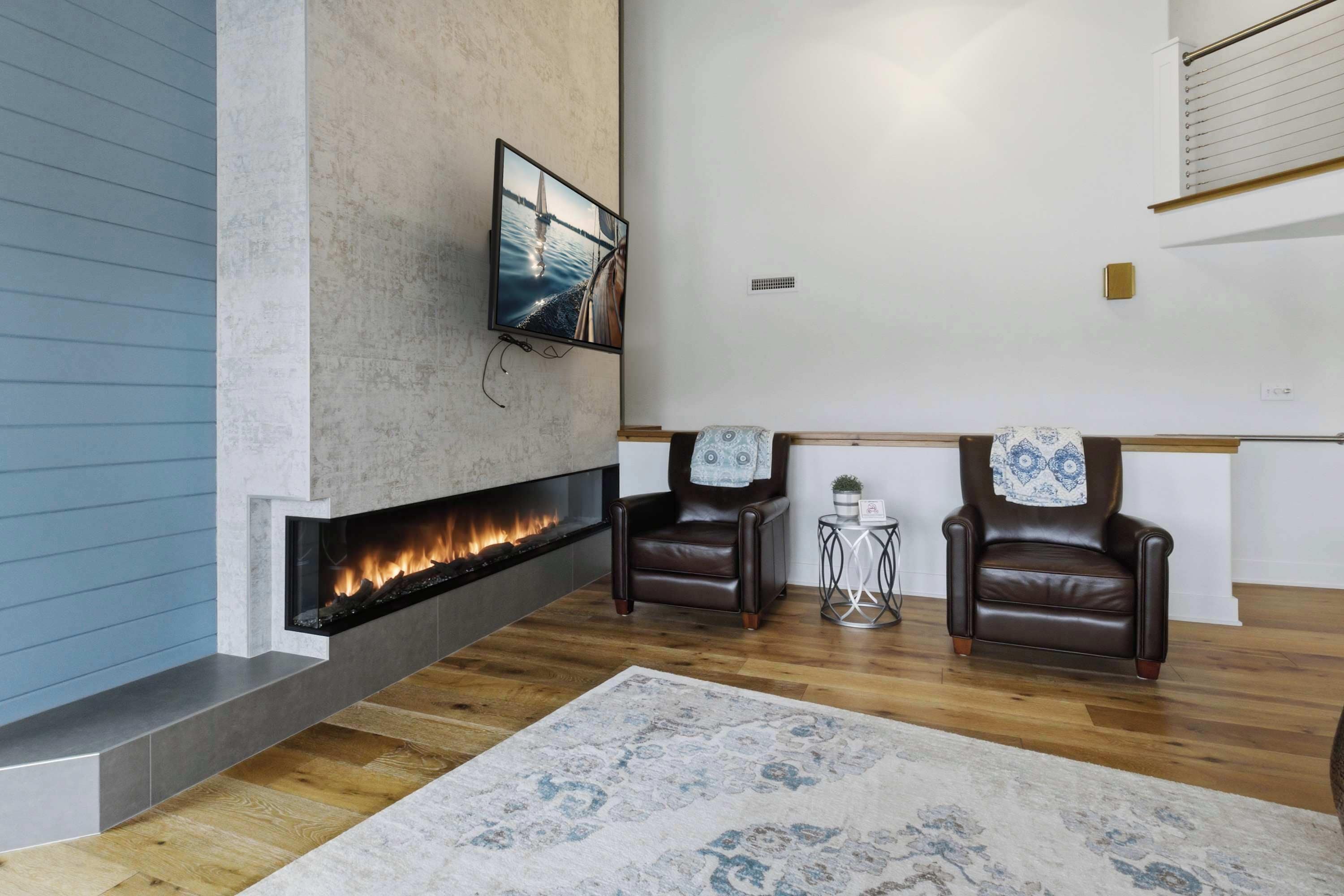 A modern living room with a fireplace, two leather chairs, a patterned rug, and a flat-screen TV mounted on the wall.