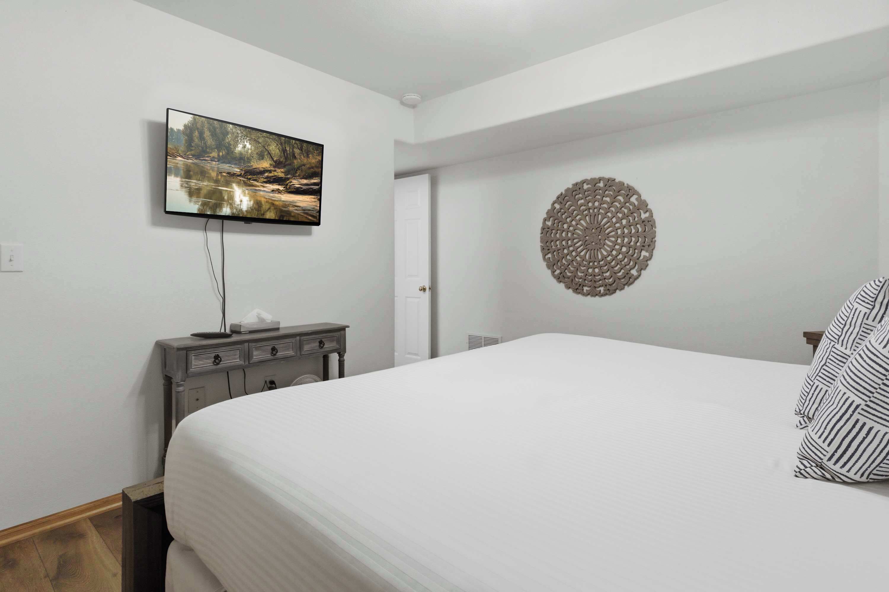 A bedroom with a bed, a flat-screen TV mounted on the wall, a bedside table with a lamp, and a decorative wall piece.