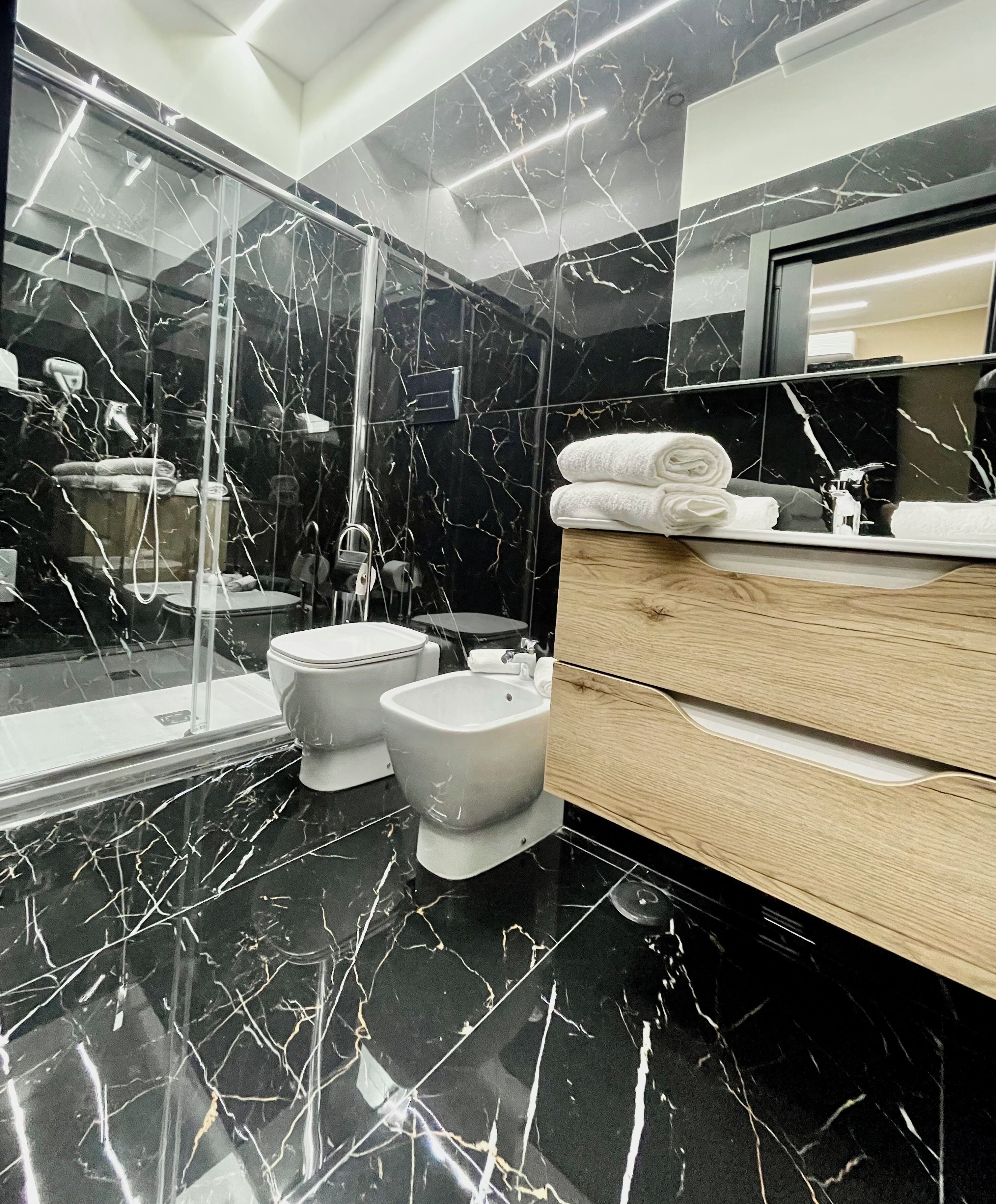 Ein modernes Badezimmer mit schwarzem Marmorboden, einer Duschkabine aus Glas, einer Toilette, einem Bidet und einem hölzernen Waschtisch mit Spiegel.