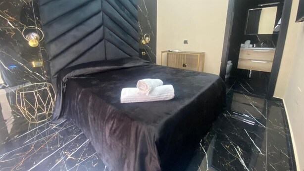 Habitación de hotel con cabecera negra, cama con colcha negra, mesita de noche, espejo y baño con lavamanos y espejo.