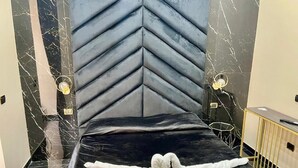 Habitación de hotel con cama negra, cabecera estampada con motivo de tablero de ajedrez y dos lámparas de noche.
