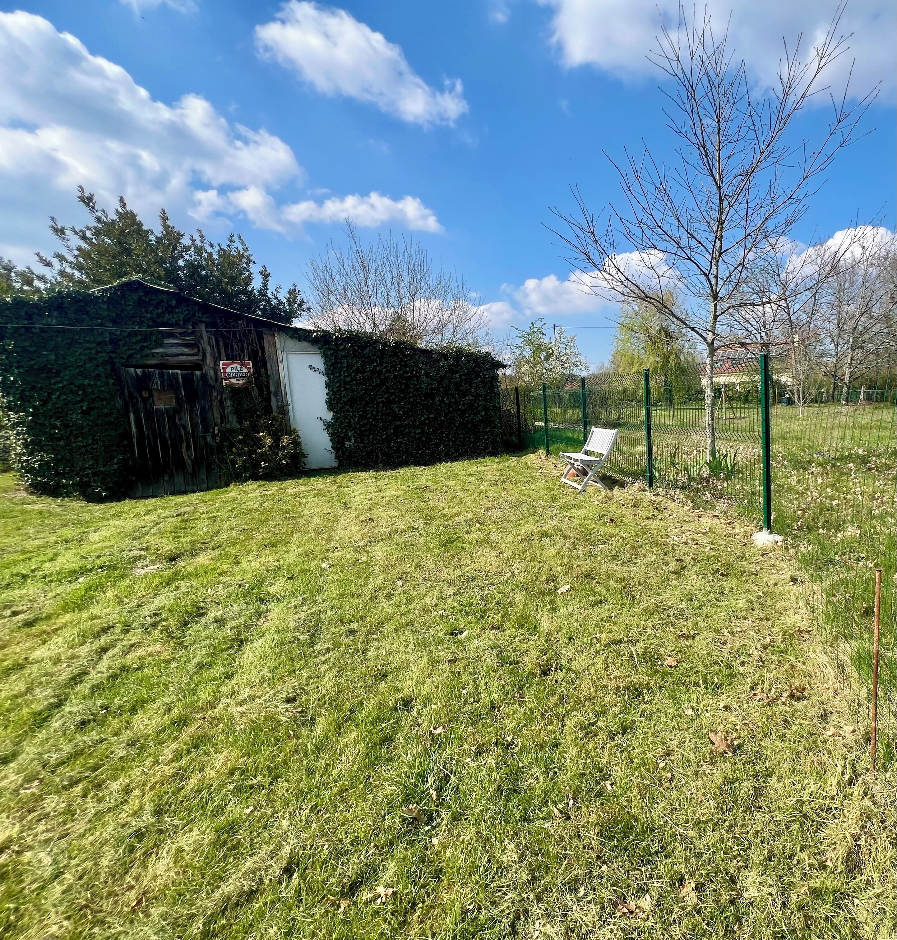 A grassy yard with a wooden shed, a white chair, and a green fence.