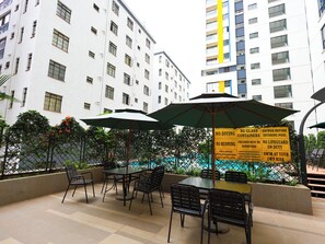 Uma área externa com mesas e cadeiras, uma piscina e um edifício com várias janelas ao fundo.