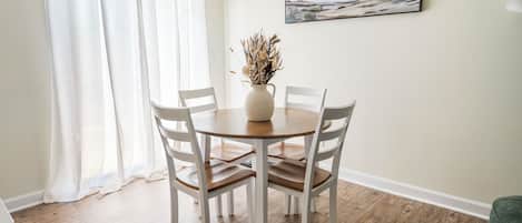 Un coin repas avec une table ronde, quatre chaises, un vase contenant des plantes séchées et un tableau accroché au mur.