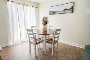 Un comedor con una mesa redonda, cuatro sillas, un florero con plantas secas y un cuadro en la pared.