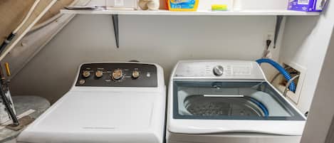 A compact laundry area with two washing machines, a shelf with cleaning supplies, and a power outlet.