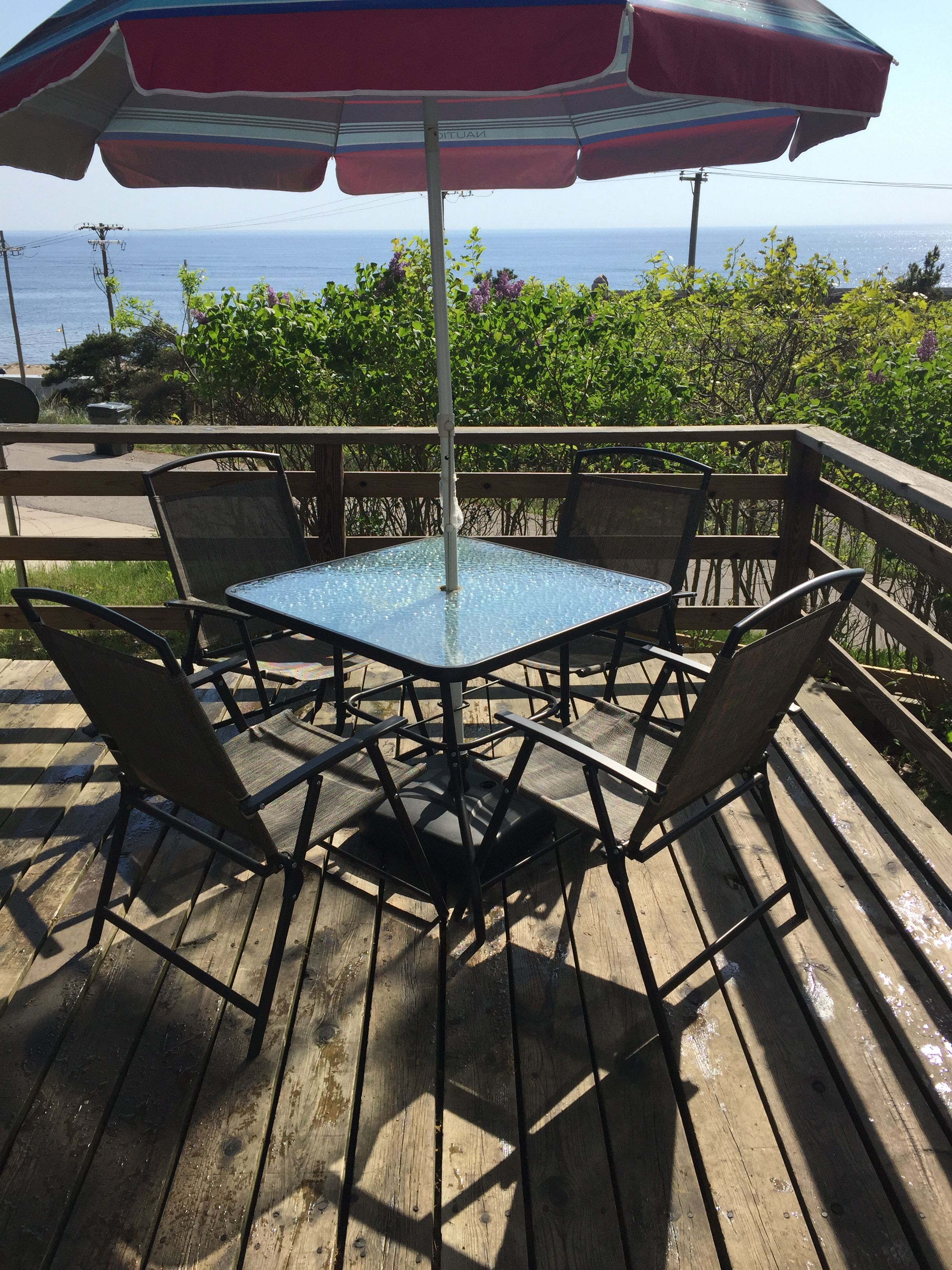 A wooden deck with a glass-top table and metal chairs, an umbrella, and a view of the ocean.