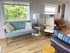 A living room with a sofa, armchairs, a small table with a lamp, and a view of greenery outside. - Hilltop Haven, Stunning Lake Michigan Views (Muskegon)