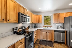 A modern kitchen with wooden cabinets, stainless steel appliances, and a window providing a view of greenery. - Big Sky Escape! 5 minute walk from Town Center! (Big Sky)