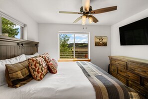 Ein Schlafzimmer mit einem Bett, einem Holz-Kopfteil, einem Deckenventilator, einem großen Fenster mit Ausblick und einem angebrachten Fernseher.