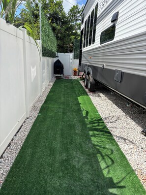 A narrow pathway with artificial green turf, flanked by a white fence and a gravel bed. A trailer is parked adjacent to the fence. - Confortable RV/Near Beaches & Downtown WPB. (West Palm Beach)
