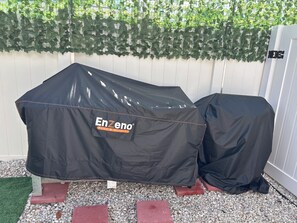 Deux housses de barbecue portant la marque « Enzeno Home & Outdoor », posées sur une surface de gravier, avec une clôture blanche et un feuillage verdoyant en arrière-plan.