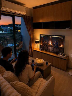 A living room with a large flat-screen TV displaying a video game, a sofa, a coffee table, and a balcony with a view of the city. - La Casita Hua Hin Premium Stay with Playstation (Tambon Hua Hin)