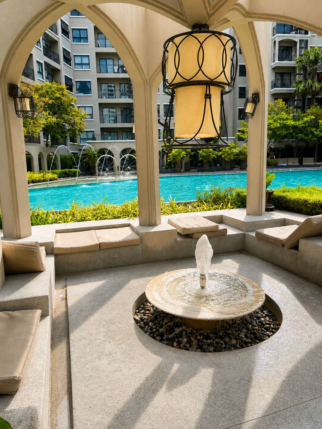 Un espace aménagé au bord de la piscine, avec une fontaine, des sièges et une vue sur le bâtiment de l’hôtel.