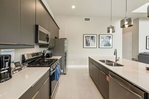 A modern kitchen with stainless steel appliances, a large island with a sink, and artwork on the walls. - 2 in 1 | 9BR for Big Family Reunions (Kissimmee)