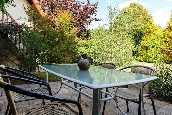An outdoor patio with a glass table and wicker chairs, surrounded by lush greenery and trees.