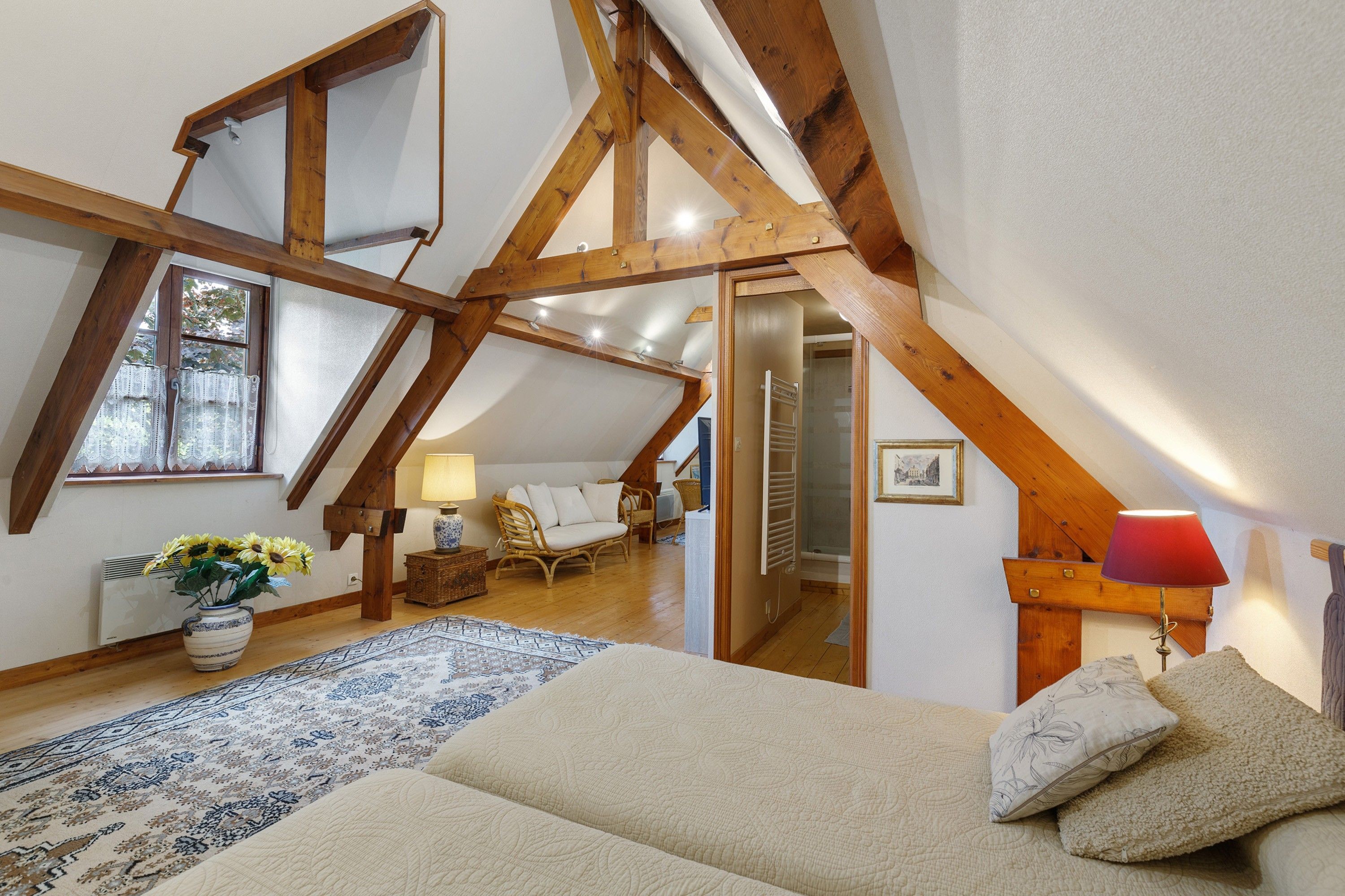 Ein Schlafzimmer mit geneigter Decke, Holzbalken, einem Bett, einem Nachttisch, einer Lampe, einem Sessel, einem Fenster mit Spitzenvorhängen und einer Vase mit Blumen.