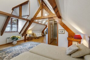 Et soveværelse med skrå væg, træbjælker, en seng, et natbord, en lampe, en stol, et vindue med blondegardiner og en vase med blomster.