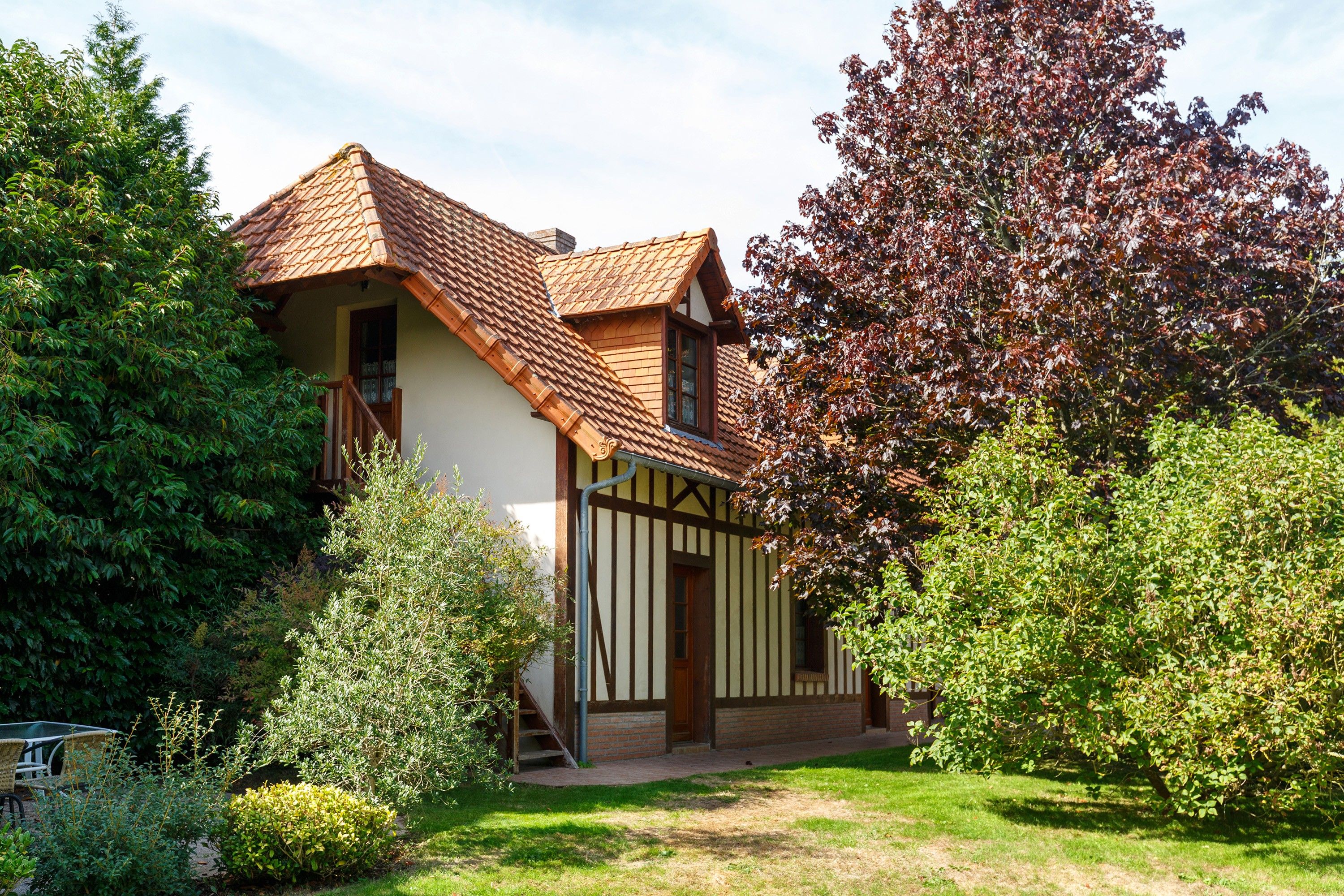 Een traditioneel huis met een schuin dak, een houten trap naar de ingang, en een verzorgde tuin met verschillende struiken en een boom.