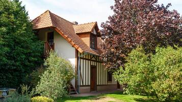 Een traditioneel huis met een schuin dak, een houten trap naar de ingang, en een verzorgde tuin met verschillende struiken en een boom.