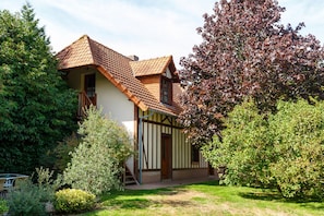 A traditional house with a sloped roof, a wooden staircase leading to the entrance, and a well-maintained garden with various shrubs and a tree.