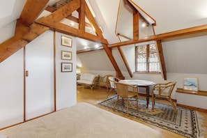 A room with a sloped ceiling, wooden beams, a dining table with chairs, a sofa, and a window with curtains.