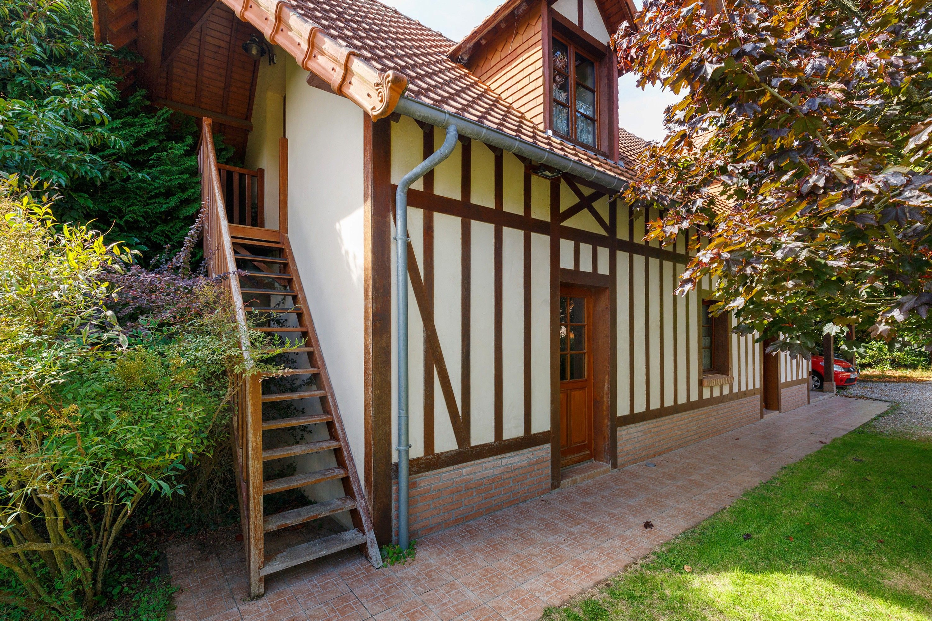 A traditional half-timbered house with a wooden staircase leading to a door, surrounded by lush greenery.