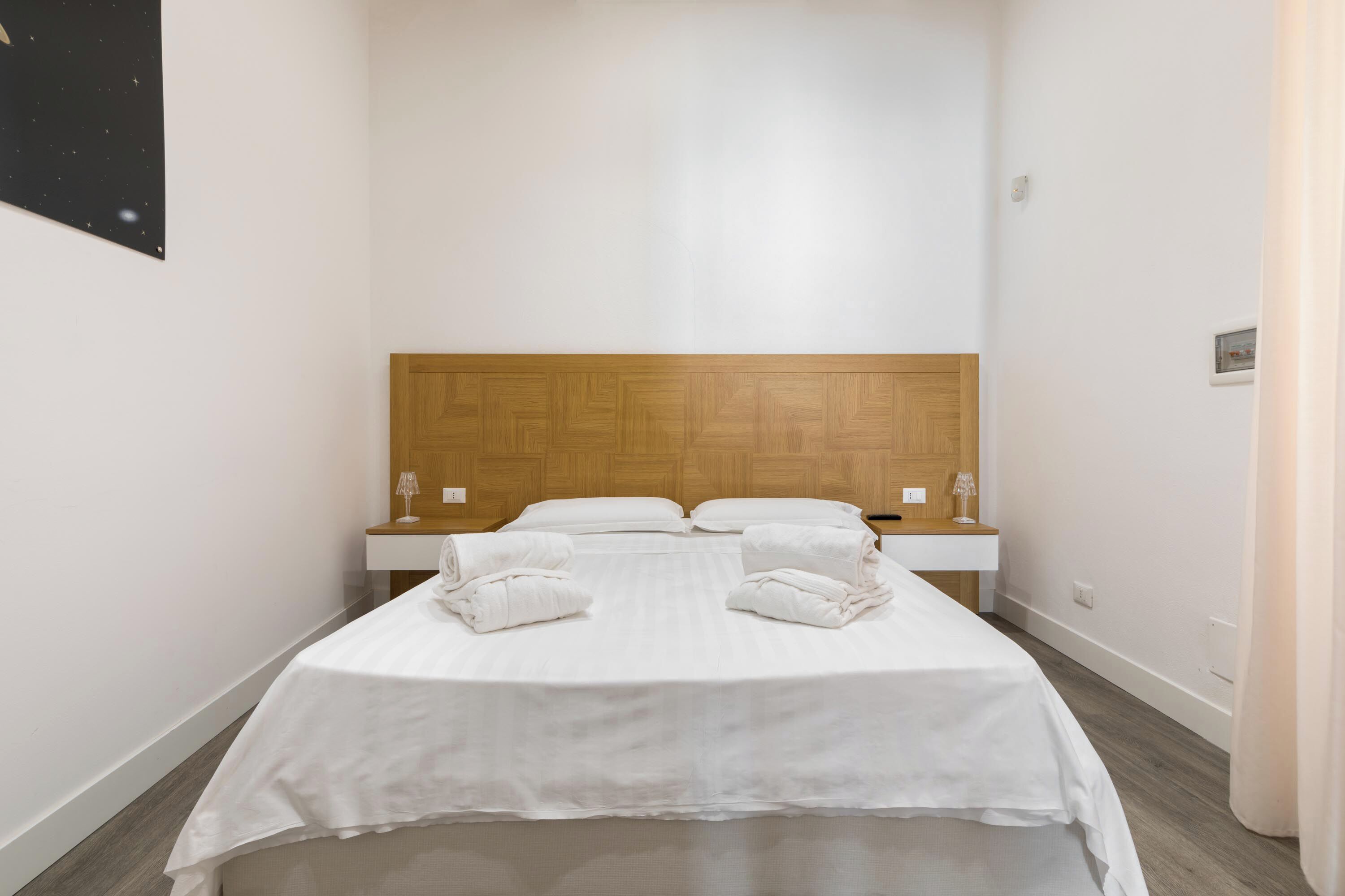 A hotel room with a wooden headboard, a bed with white linens, two bedside tables, and a nightstand.