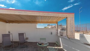 Una terraza en la azotea con una pérgola de madera, dos sillones plegables, una mesita redonda con una cesta y vasos, y vistas a edificios al fondo.