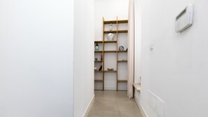 Un pasillo estrecho con una estantería de madera contra la pared, una puerta blanca al final y una pared blanca con un interruptor de luz.
