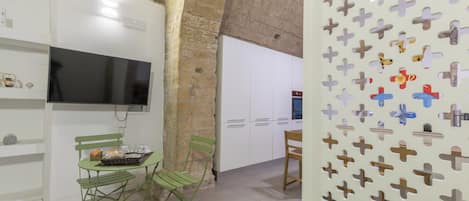 A room with a television, a dining table with chairs, a kitchen area, and a decorative wall with cut-out shapes.