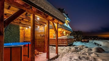 A wooden cabin with a covered outdoor seating area and a snow-covered landscape at night.