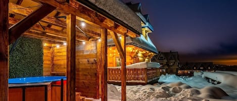 A wooden cabin with a covered outdoor seating area and a snow-covered landscape at night.