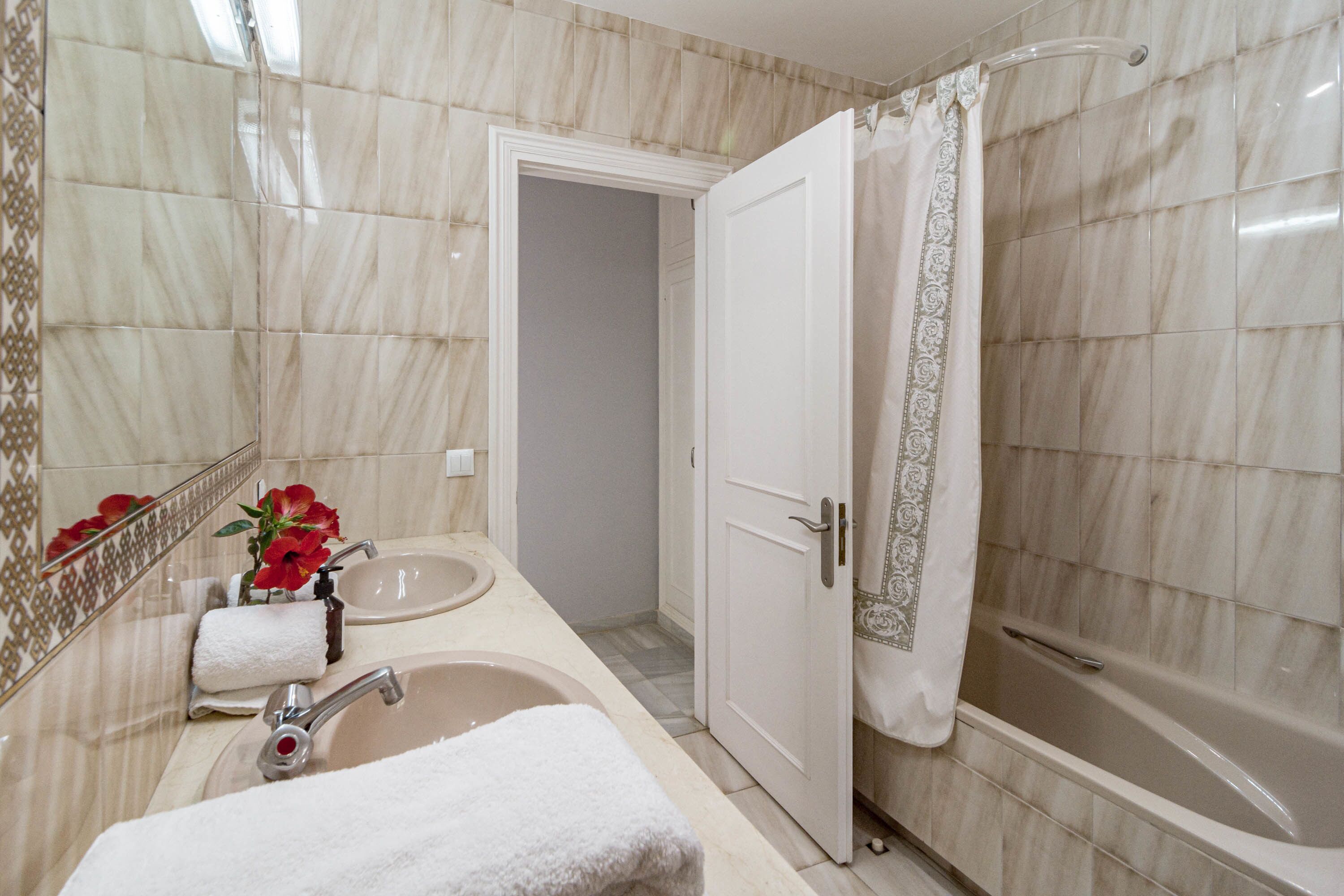 A bathroom with a marble-tiled wall, a sink with a mirror, a shower with a curtain, and a door leading to another room.