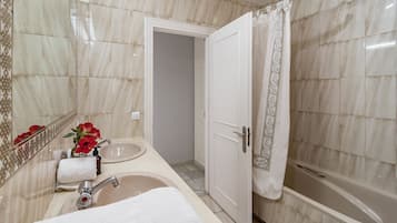 A bathroom with a marble-tiled wall, a sink with a mirror, a shower with a curtain, and a door leading to another room.