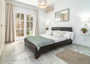 A bedroom with a bed, a window with curtains, a mirror, a wall-mounted air conditioner, and a bedside lamp.