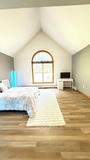 A bedroom with a sloped ceiling, a large arched window, a bed with a white and floral bedspread, a white desk with a flat-screen TV, and a white rug on the wooden floor.