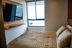 Un dormitorio con cama, televisor montado en la pared, ventana con vistas a edificios y mesita de noche con control remoto.