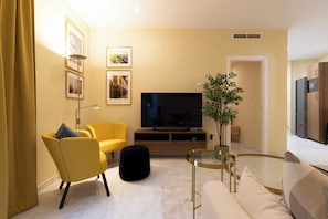Een moderne woonkamer met een flatscreen televisie, gele fauteuils, een glazen salontafel en een potplant.