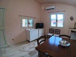 A room with a dining table, chairs, a TV, a small cabinet, and two windows. - Casa Aura Mare, Orosei (Sardegna)