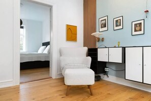 A room with a bed, a white armchair, a cabinet, and a wall with framed pictures.