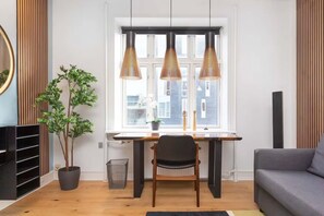 A modern living room with a wooden desk, a black chair, a grey sofa, and a large window with three pendant lights.