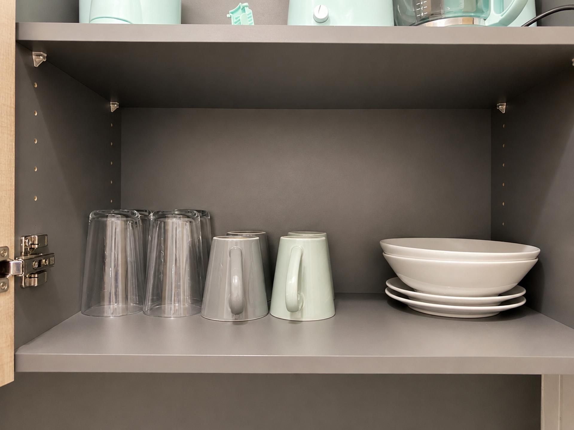 A kitchen cabinet with a grey interior, containing a stack of clear glasses, a white mug, and a white bowl.