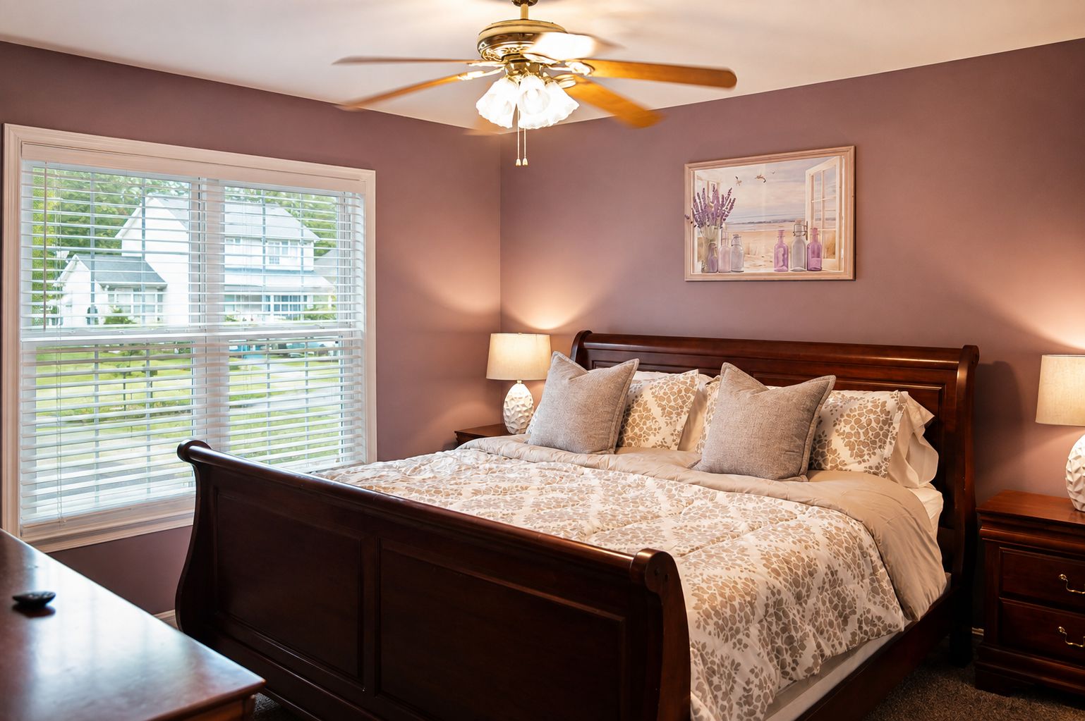A bedroom with a bed, bedside lamps, a ceiling fan, a window with blinds, and a painting on the wall.